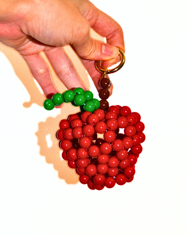 Hand holding a keychain shaped like a red apple with green leaves on a white background