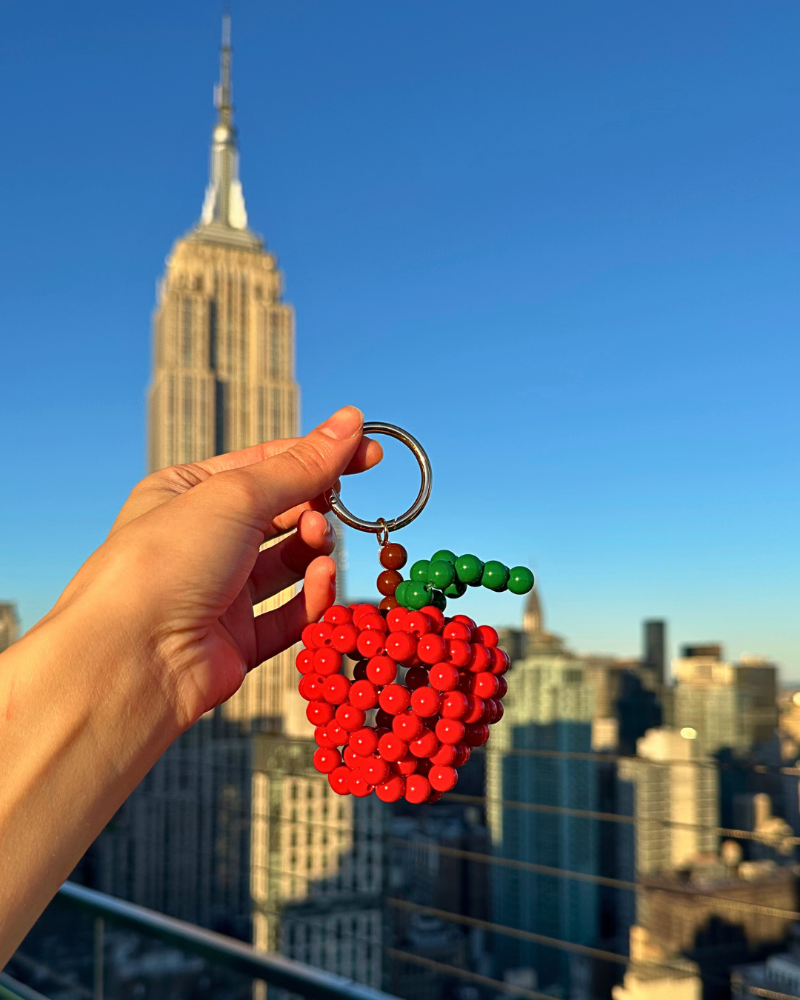 Hand holding a red beaded apple keychain with a city skyline in the background