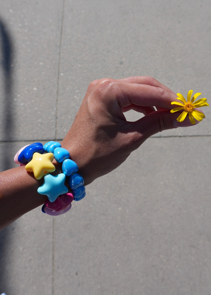 A close-up of a person's hand holding a small yellow daisy flower, wearing a stack of colorful beaded bracelets with star and heart charms in blue, yellow, and pink, set against a gray concrete background.