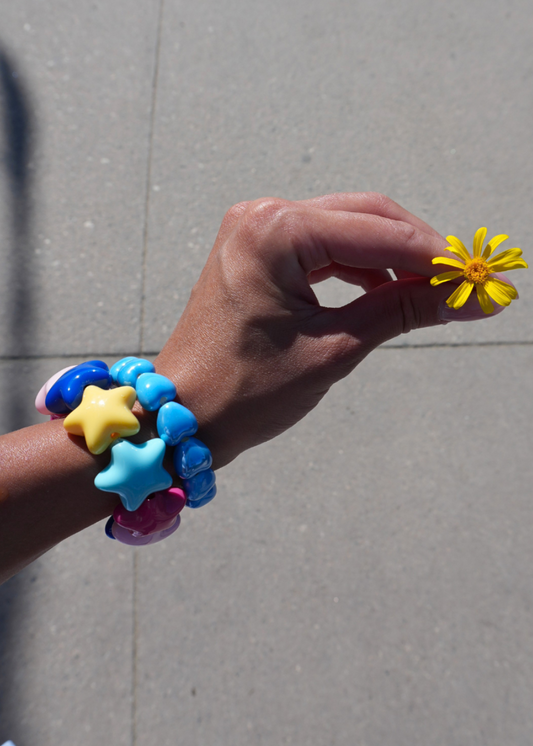 A close-up of a person's hand holding a small yellow daisy flower, wearing a stack of colorful beaded bracelets with star and heart charms in blue, yellow, and pink, set against a gray concrete background.