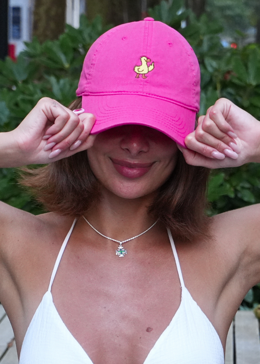 A woman wearing a bright pink baseball cap with a small yellow duck logo on the front, posing outdoors with her hands on the brim. She is wearing a white halter top and a silver necklace with a green pendant, with lush green foliage in the background.